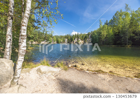 The Piskovna lake in Adrspach-Teplice Rocks area, in Hradec Kralove Region in Czech Republic, Europe. Scenic lake surrounded by lush green forest, with birch trees in the foreground under sunny sky 130453679
