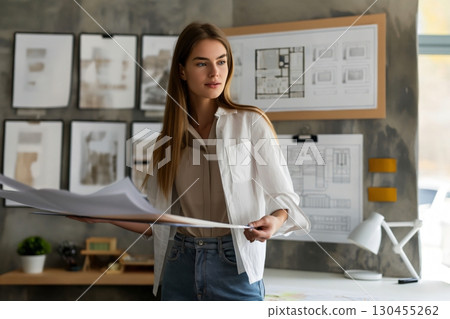 Young female architect is reviewing blueprints in her modern office, surrounded by design plans and architectural drawings 130455262