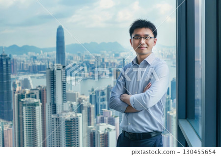 Young businessman is posing with his arms crossed in a modern office overlooking a vibrant city 130455654