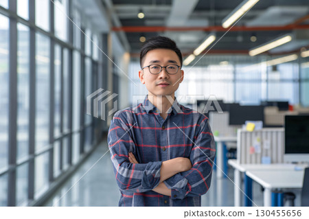 Young asian businessman is standing in a modern office with his arms crossed, looking serious and confident Young asian businessman is standing in a modern office with his arms crossed, looking serious and confident 130455656