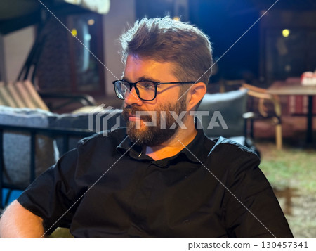 Bearded man wearing glasses and black shirt seated outdoors in the evening. Portrait, lifestyle, leisure, hospitality, social atmosphere and night terrace environment. 130457341