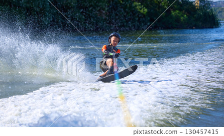 Boy in black helmet leaning forward while water skiing with splash trail Boy in black helmet leaning forward while water skiing with splash trail 130457415