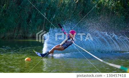 Girl in pink helmet leaning dramatically while water skiing on lake surface 130457418
