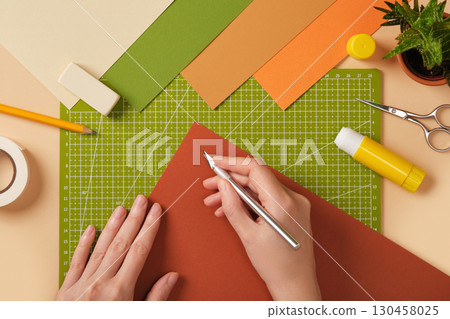 Hands holding knife and brown paper sheet on cutting mat with stationery around. Crafting process. Hands holding knife and brown paper sheet on cutting mat with stationery around. Crafting process. 130458025