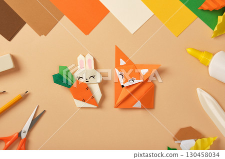 Paper rabbit with carrot and fox origami figures on desk with colored sheets, scissors, pencil, eraser and glue. Craft toy for kids. Paper rabbit with carrot and fox origami figures on desk with colored sheets, scissors, pencil, eraser and glue. Craft toy for kids. 130458034