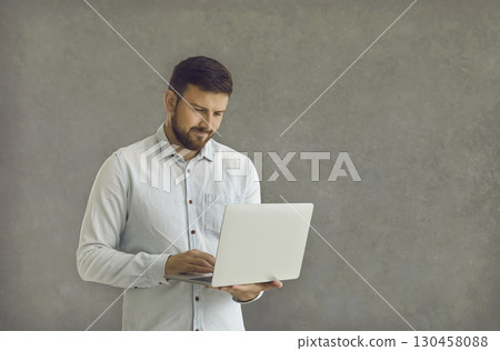 Young and confident man standing on a gray background with a laptop in his hands. Young and confident man standing on a gray background with a laptop in his hands. 130458088