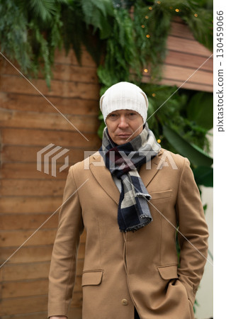 Winter portrait of caucasian man wearing white beanie and cozy scarf, with blurred Christmas festive background. Cold winter weather. Outdoors. Xmas holiday. 130459066
