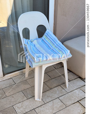 Plastic chair on a balcony with striped towel folded in morning light. Leisure, vacation, simplicity and everyday comfort during seasonal resort travel and accommodation. Plastic chair on a balcony with striped towel folded in morning light. Leisure, vacation, simplicity and everyday comfort during seasonal resort travel and accommodation. 130459667