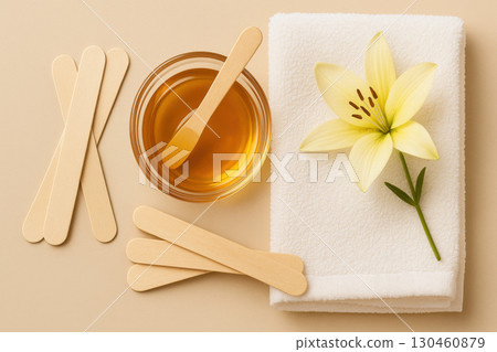 Spa waxing setup with honey wax in glass bowl, wooden spatulas, flower sprig, and soft towel on beige background, promoting beauty and self care 130460879