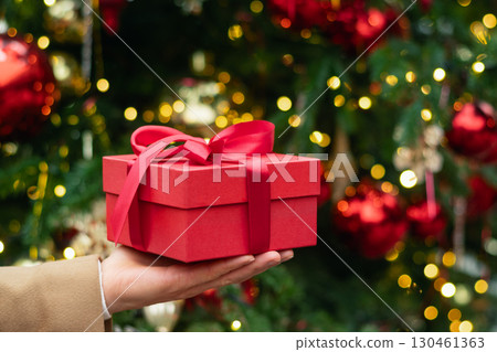 Man hand holding red gift box or present against Christmas tree. Xmas shopping. Close up. Man hand holding red gift box or present against Christmas tree. Xmas shopping. Close up. 130461363
