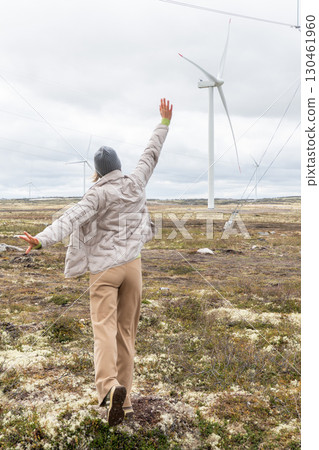 Woman in beige pants and a gray jacket raises her arms joyfully near wind turbines and power lines in Teriberka, Russia 130461960