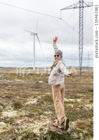 Woman in beige pants and a gray jacket raises her arms joyfully near wind turbines and power lines in Teriberka, Russia Woman in beige pants and a gray jacket raises her arms joyfully near wind turbines and power lines in Teriberka, Russia 130461961
