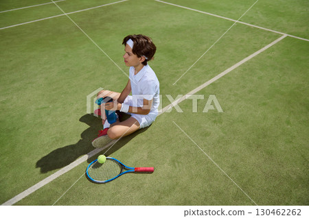 Young Tennis Player Resting on Court Holding Water Bottle and Tennis Gear 130462262