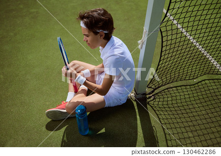 Focused Young Tennis Player Sitting on Court by the Net Outdoors Focused Young Tennis Player Sitting on Court by the Net Outdoors 130462286