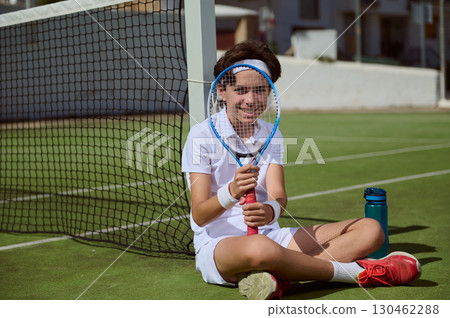 Young Boy Resting with Tennis Racket on Sunny Court 130462288
