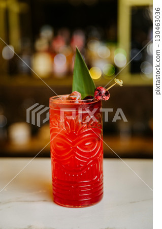 Tropical red cocktail with cherries and leaf garnish in a tiki glass on a bar counter 130463036