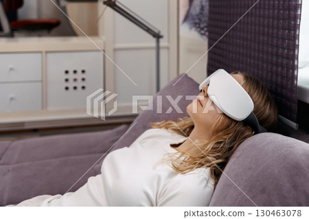 Eye massage device. Young girl wearing glasses for massage relaxing on sofa at home. Woman takes care of her health and skin care 130463078