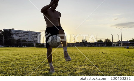 Young man juggling soccer ball on stadium at sunset. Professional footballer kicking ball at green field. Sportsman practicing tricks at meadow with sunlight at background. Freestyle football 130463221