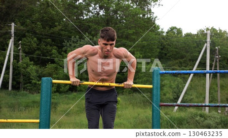 Hardy shirtless sportsman doing push ups on horizontal bar at nature. Strong and muscular guy training at sports ground. Young athletic guy exercising outdoor. Concept of active and healthy lifestyle 130463235