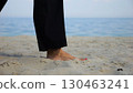 Legs of adult woman stepping along sea shore. Female feet walking on sand at the beach with seascape at background. Barefoot lady strolling near ocean coast. Concept of summer vacation. 130463241