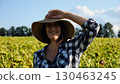 Happy smiling female farmer looks into camera standing near sunflower field. Portrait of adult beautiful agronomist in straw hat with yellow plantation at background. Agricultural business concept 130463245