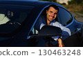 Young smiling entrepreneur in shirt leaning out of driver window at modern electric car and looking into camera. Portrait of handsome successful businessman in new electrical vehicle. Slow mo 130463248