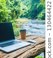 Laptop and coffee cup on rustic table by river, surrounded by lush greenery 130464422