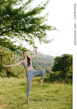 Young athletic woman practicing yoga outdoors in a serene green landscape, embodying tranquility and strength with a smile Young athletic woman practicing yoga outdoors in a serene green landscape, embodying tranquility and strength with a smile 130464448