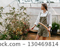 Young woman in a greenhouse wearing a white shirt and green overalls, tending to plants with a hose, expressing joy and connection to nature 130469814