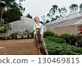 Young woman holding a potted plant in a greenhouse, showcasing joy and connection to nature with a casual outfit and lush greenery in the background 130469815