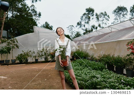 Young woman holding a potted plant in a greenhouse, showcasing joy and connection to nature with a casual outfit and lush greenery in the background Young woman holding a potted plant in a greenhouse, showcasing joy and connection to nature with a casual outfit and lush greenery in the background 130469815