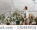 Young woman gardening in a greenhouse, wearing a casual outfit, showcasing the joy of nurturing plants in a vibrant, green environment 130469818