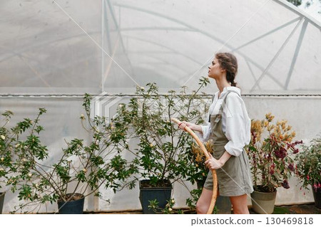 Young woman gardening in a greenhouse, wearing a casual outfit, showcasing the joy of nurturing plants in a vibrant, green environment 130469818