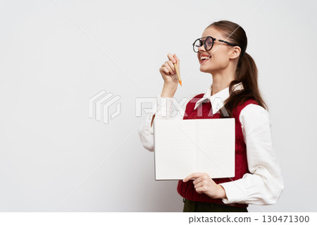 Smiling young woman holding a blank notebook and pencil, wearing glasses, dressed in a stylish red vest against a neutral background, representing creativity and education Smiling young woman holding a blank notebook and pencil, wearing glasses, dressed in a stylish red vest against a neutral background, representing creativity and education 130471300