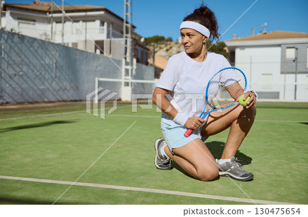 Confident Woman Kneeling on Tennis Court Ready for an Exciting Match 130475654