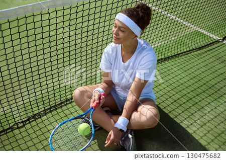 Woman Sitting by Tennis Court Net Holding Racket 130475682