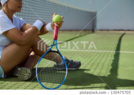 Tennis Player Sitting on Court Holding Ball and Racket in Relaxing Moment 130475705
