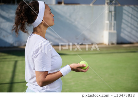 Focused Female Tennis Player Preparing for a Game on the Court. A woman dressed in sports attire holds a tennis ball, ready to begin a game on a sunny court. Focused Female Tennis Player Preparing for a Game on the Court. A woman dressed in sports attire holds a tennis ball, ready to begin a game on a sunny court. 130475938