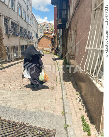 Elderly woman in black burqa with grocery bags walking up cobbled city street Elderly woman in black burqa with grocery bags walking up cobbled city street 130477035