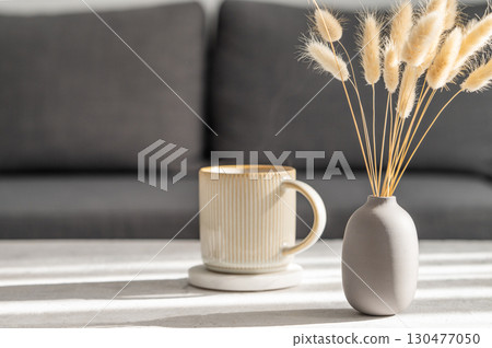 Beige ceramic cup of coffee with steam on a light table with soft shadows, vase and notebook Beige ceramic cup of coffee with steam on a light table with soft shadows, vase and notebook 130477050