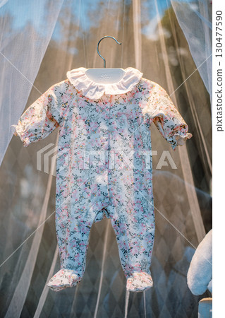 Floral baby romper hanging on a white hanger inside a softly lit display with sheer curtains and plush toys. Vintage childhood, boutique nursery style, delicate textures, early parenthood 130477590