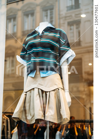 Mannequin dressed in layered striped polo, button-down shirt, and pleated beige skirt in boutique window. Gender-fluid softwear, comfort silhouettes, unstructured design, inclusive wardrobe Mannequin dressed in layered striped polo, button-down shirt, and pleated beige skirt in boutique window. Gender-fluid softwear, comfort silhouettes, unstructured design, inclusive wardrobe 130477601