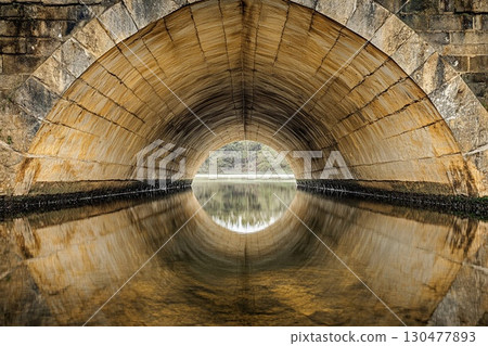 Vaulted bridge structure creates stunning mirror reflection beneath in calm water during early morning light 130477893