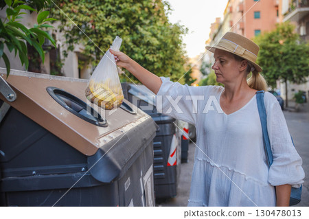 Middle-aged woman in a straw hat and white dress disposes of organic waste into a recycling container on a residential street in Rome. 130478013