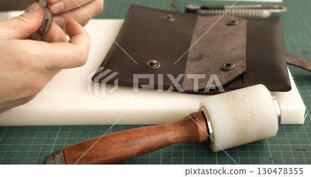 Leatherworker preparing brown leather wallet with pricking iron and mallet closeup 130478355
