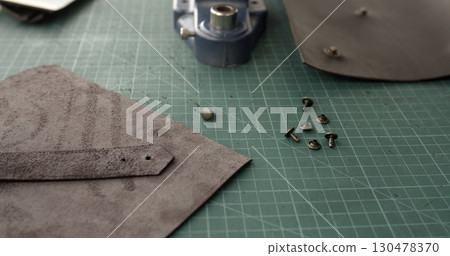 Leather pieces with metal rivets and press tool on green cutting mat in craftsman workshop closeup Leather pieces with metal rivets and press tool on green cutting mat in craftsman workshop closeup 130478370