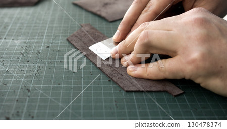 Craftsman skiving surface of leather strip with blade tool on green cutting mat in workshop Craftsman skiving surface of leather strip with blade tool on green cutting mat in workshop 130478374