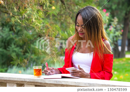 African woman in red attire enjoys a drink while writing in a notebook outdoors 130478599