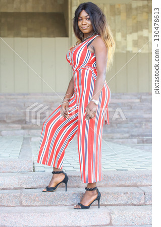 An African woman stands gracefully on steps, showcasing a stylish striped jumpsuit 130478613