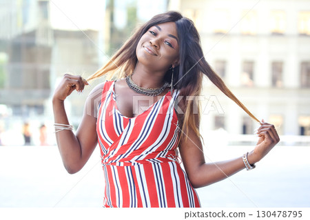Beautiful African woman posing outdoors while playing with her hair in a stylish outfit 130478795